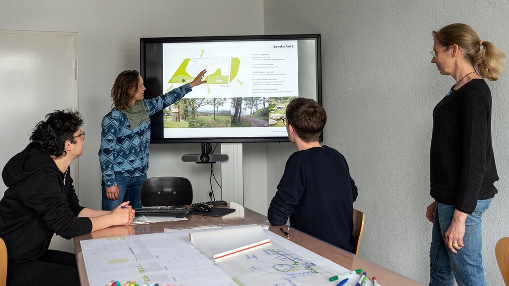 In einem Besprechungsraum steht eine Frau vor einem Smartboard und erläutert drei vor ihr sitzendeden Personen etwas darauf.