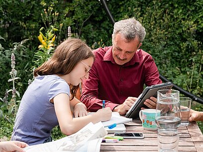 Ein Vater sitzt mit seiner Tochter im Garten. Sie schreibt auf einen Block, er schaut ihr zu.