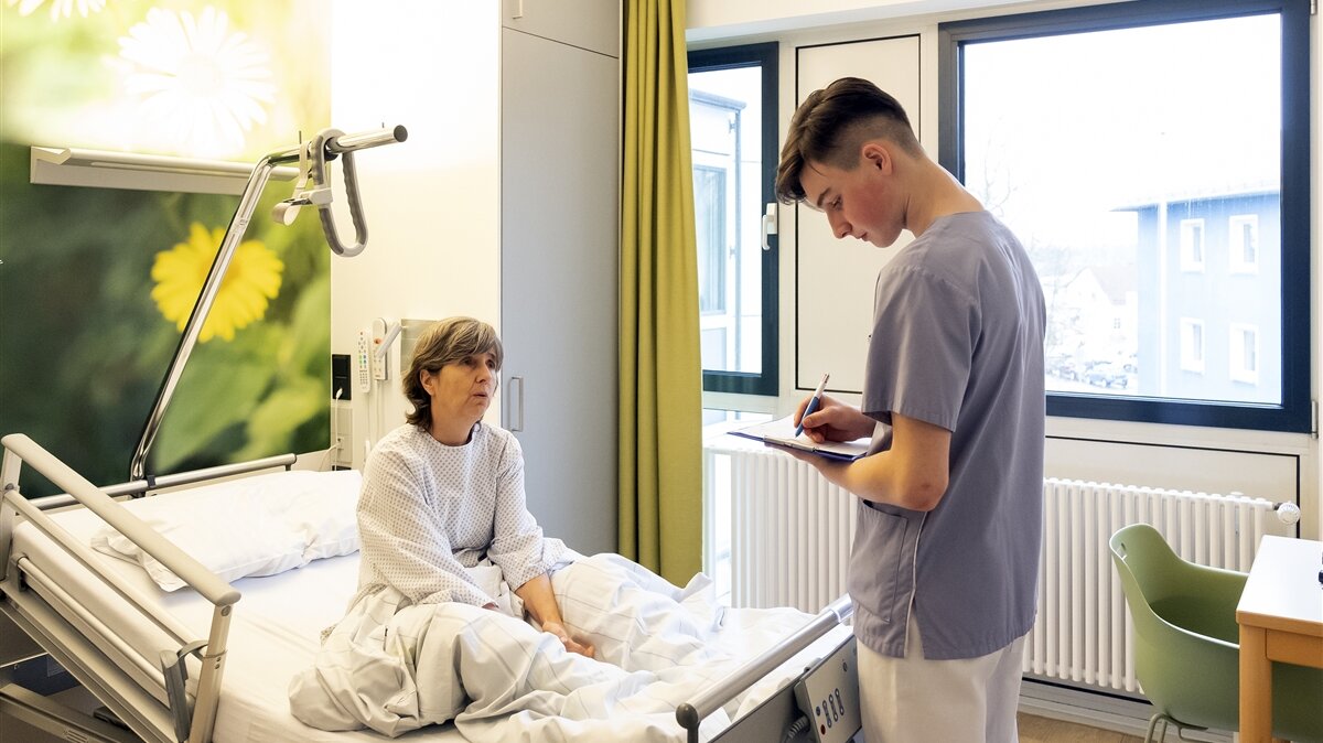 Ein junger Mann notiert etwas am Krankenbett einer Patientin.