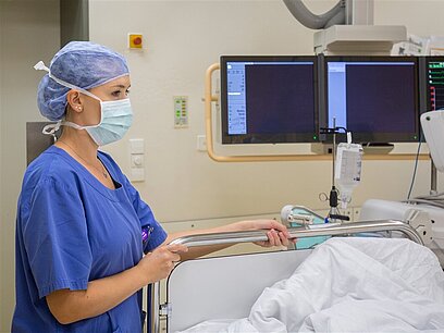 Eine Frau mit Mundschutz und Haarnetz schiebt ein Krankenbett in einen Untersuchungsraum.