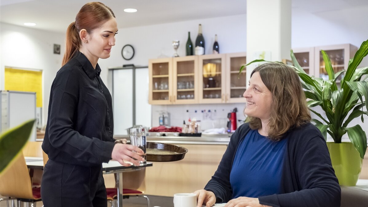 Andreea bringt einem Gast ein Getränk an den Tisch.