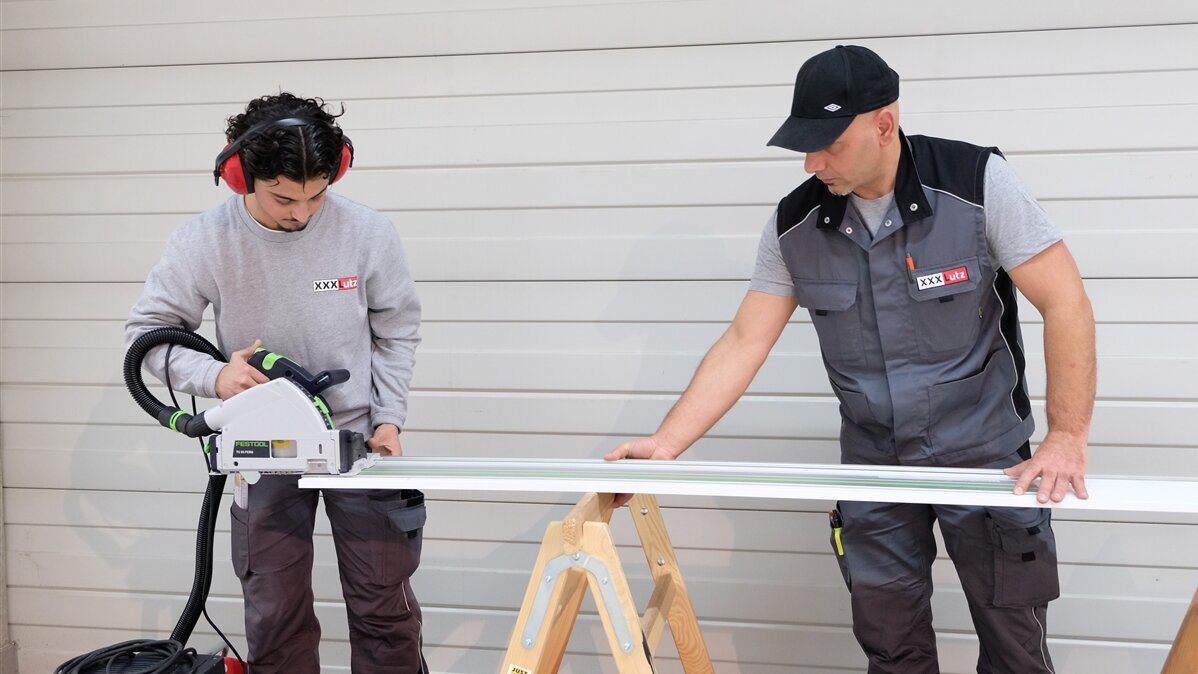Zainal passt die Länge eines Regalbretts mit einer Tischkreissäge an, während sein Kollege das Brett festhält.