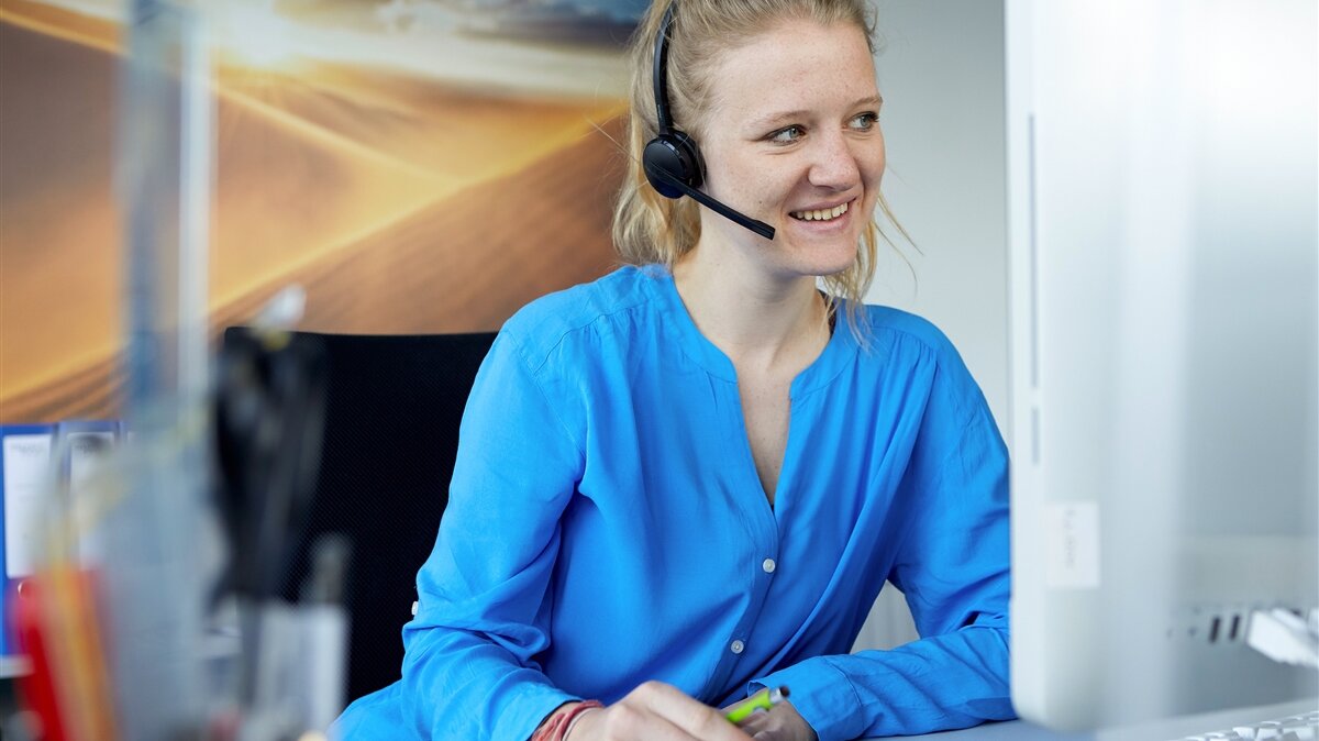 Eine junge Frau mit Headset sitzt an ihrem Schreibtisch und lächelt.