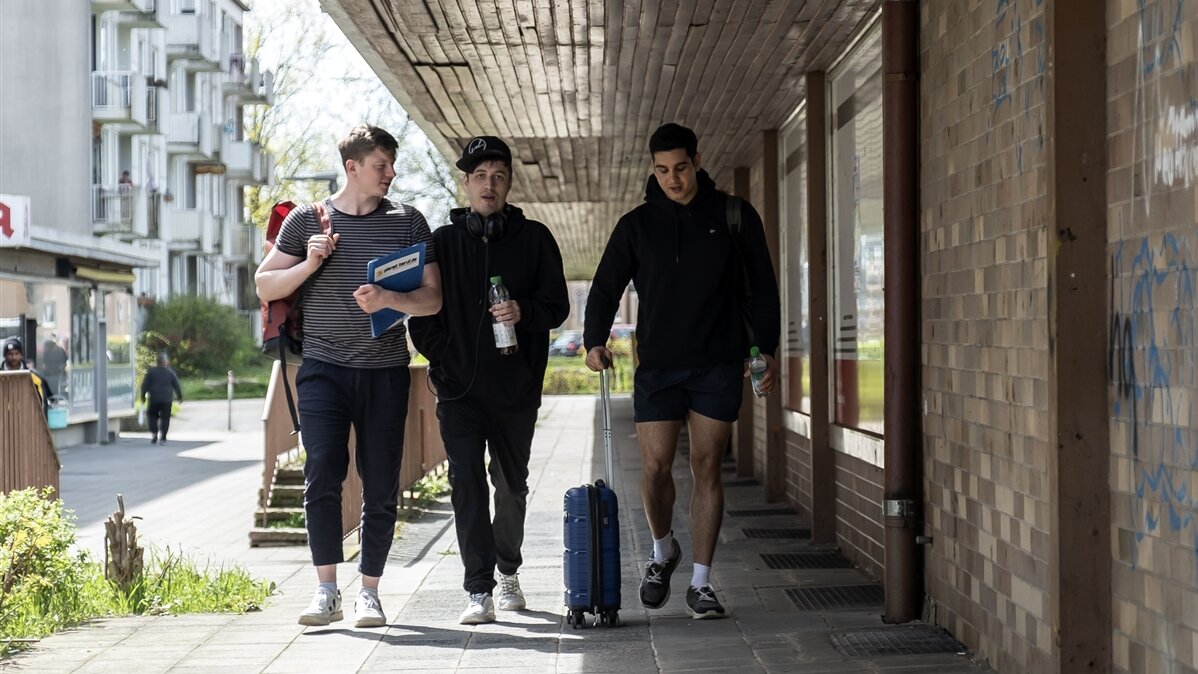 Drei junge Männer, einer mit Koffer, laufen auf der Straße. 