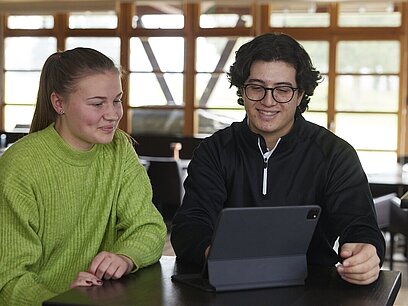 Zwei junge Menschen sitzen vor einem Tablet und unterhalten sich.