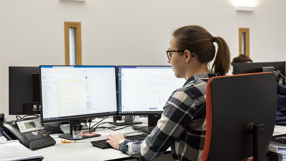 Eine junge Frau mit Brille sitzt vor zwei Computermonitoren und tippt Daten über eine Tastatur ein.