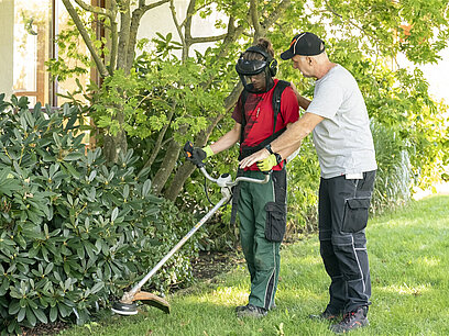 Ein Mann in Arbeitskleidung zeigt einem jungen Mann in Schutzkleidung, wie ein Rasentrimmer verwendet wird.