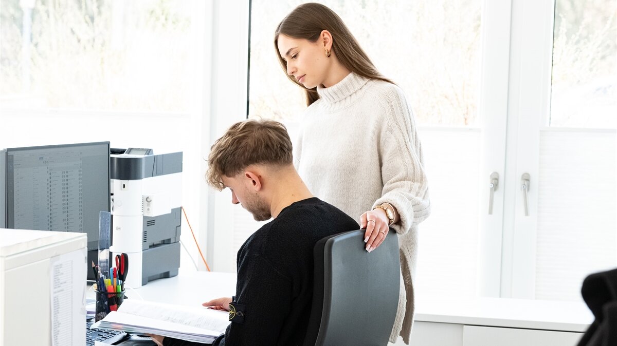 Ein junger Mann und eine junge Frau schauen sich vor einem PC im Büro etwas in einer Akte an.