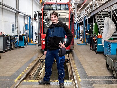 Ein junger Mann in Arbeitskleidung steht in einer Werkhalle vor einer Straßenbahn.