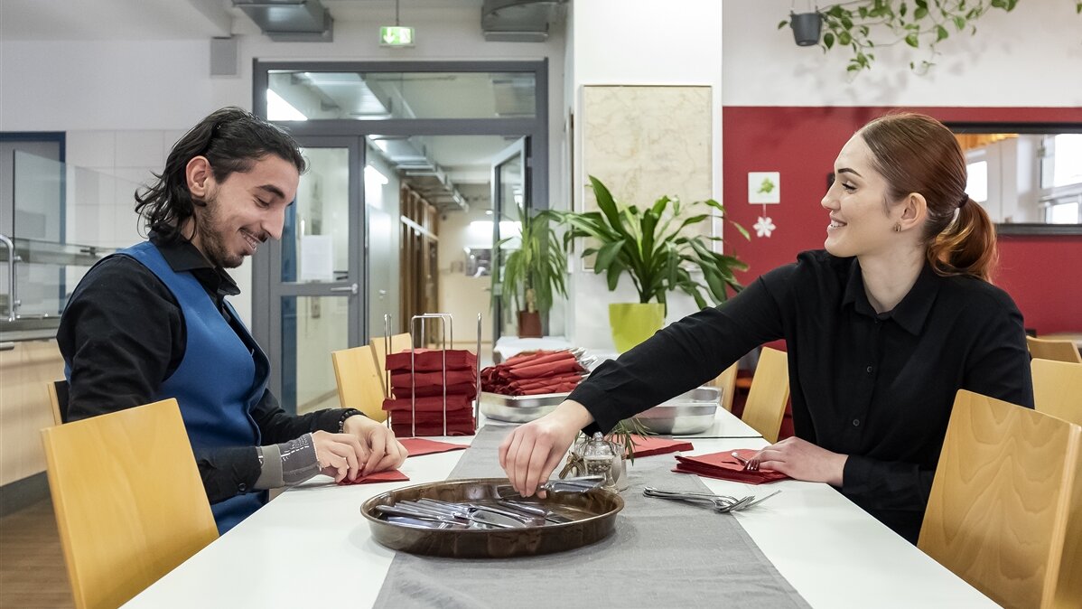 Andreea und ihr Kollege wickeln Besteck in Servietten ein.