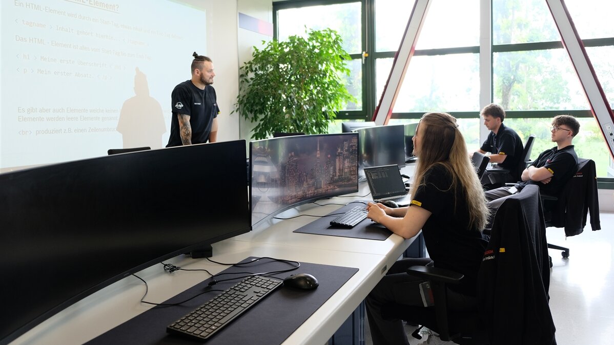 Ayliah und ihre Kollegen sitzen vor Computern, während ihr Ausbilder vor einem Whiteboard steht.