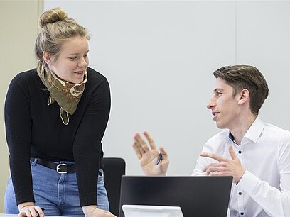 Ein junger Mann sitz vor einem Laptop und spricht mit einer neben ihm stehenden jungen Frau.