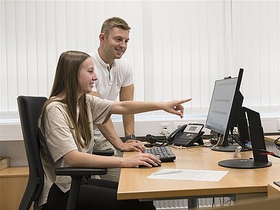 Ein Mann steht neben einer jungen Frau, die vor einem Computer sitzt und auf den Bildschirm zeigt.
