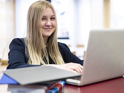 Eine junge Frau am Laptop, die in die Kamera lächelt.
