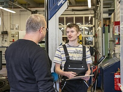 Ein junger Mann mit Klemmbrett in der Hand berät einen Mann in einer Werkstatt.