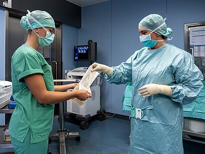 Zwei junge Frauen stehen mit Kittel und Mundschutz in einem Operationssaal und reichen sich Handschuhe.
