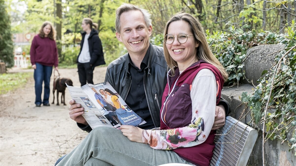 Ein Mann und eine Frau sitzen zusammen auf einer Bank und haben das Elternmagazin in der Hand. 
