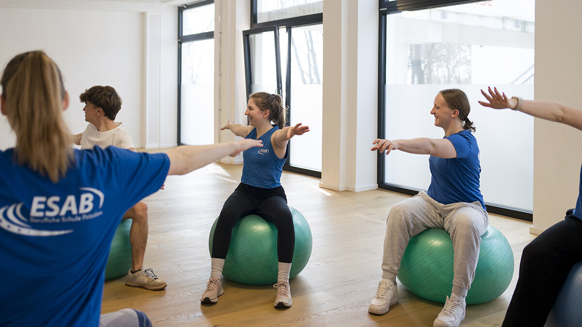 Liselotte sitzt auf einem Pezziball in der Mitte der Klasse und führt eine sportliche Übung aus.