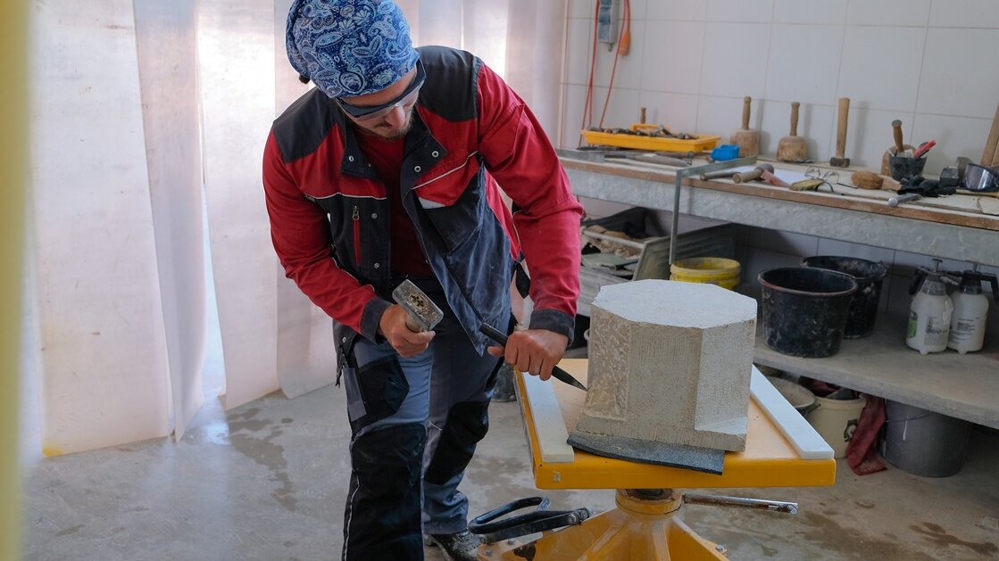 Leonhard arbeitet in der Werkstatt mit Fäustel und Spitzeisen den Sockel an einem Steinblock heraus.