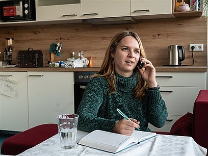 Eine Jugendliche sitzt in einer Küche, telefoniert mit dem Handy und macht sich Notizen.