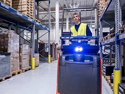 Ein junger Mann in einer Signalweste fährt mit einem Gabelstapler durch einen Gang in einer Lagerhalle.