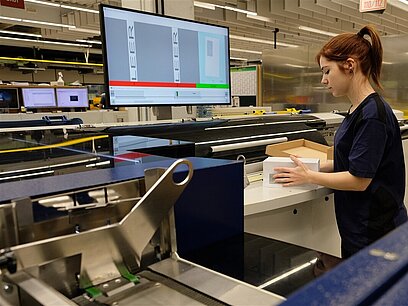 Eine Frau steht vor einer Maschine und einem Bildschirm und verpackt Umschläge.