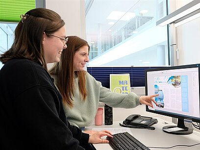 Zwei junge Frauen sitzen vor dem Computer und bearbeiten einen Flyer.