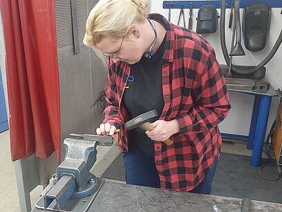 Eine junge Frau bearbeitet Werkstück aus Metall, das sie in einen Schraubstock eingespannt hat.