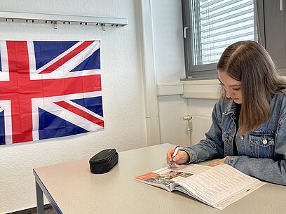 Melina sitzt an einem Tisch vor einer Flagge Großbritanniens und arbeitet in einem Heft.