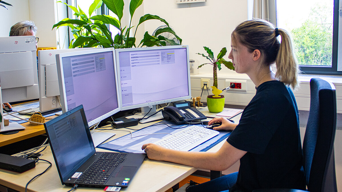 Alicia arbeitet am Schreibtisch im Büro an einem Laptop und zwei zusätzlichen Bildschirmen.