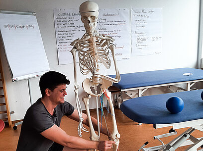 Ein junger Mann kniet neben einem künstlichen Skelett und betrachtet dessen Aufbau.