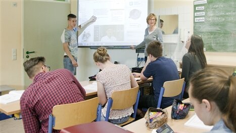 Ein Schüler und eine Lehrerin stehen an der Leinwand vor Schülern in einem Klassenzimmer.