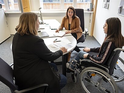 Eine Berufsberaterin im Gespräch mit einer Mutter und deren Tochter, die im Rollstuhl sitzt.