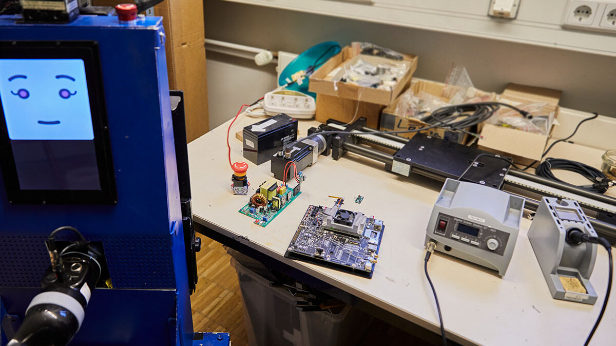 Ein Roboter mit Smiley auf seinem Monitor vor einem Tisch mit elektronischen Geräten