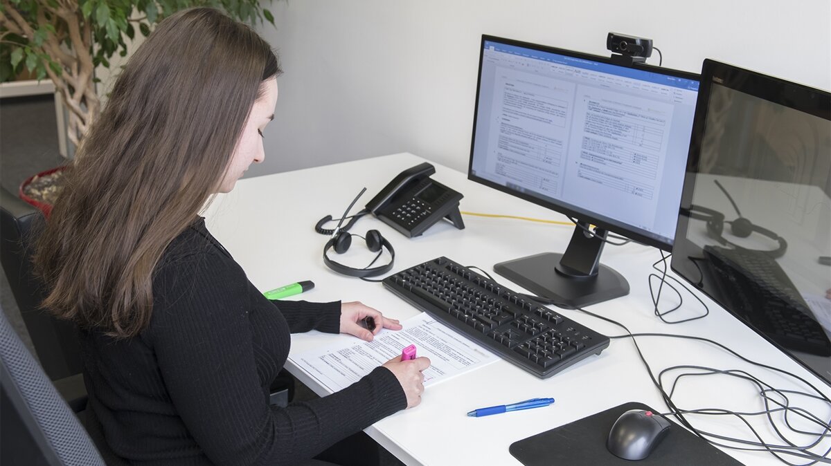 Eine junge Frau gleicht ein Dokument mit einer Vorgabe am Computer ab.