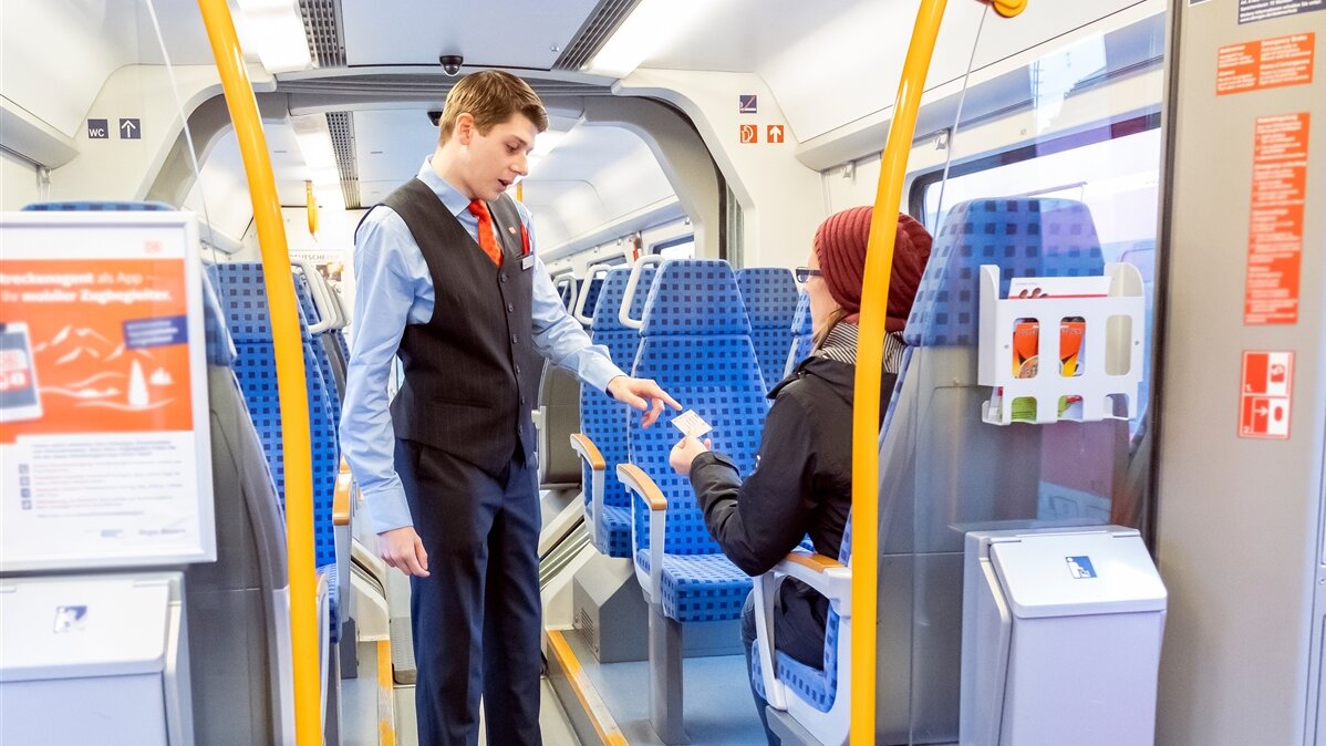 In einem Zug wird die Fahrkarte einer Reisenden von einem jungen Mann in Uniform kontrolliert.