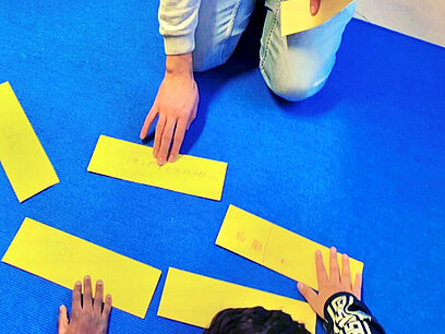 Christoph legt für eine Übung mit zwei Kindern gelbe Zettel auf eine Matte am Boden.