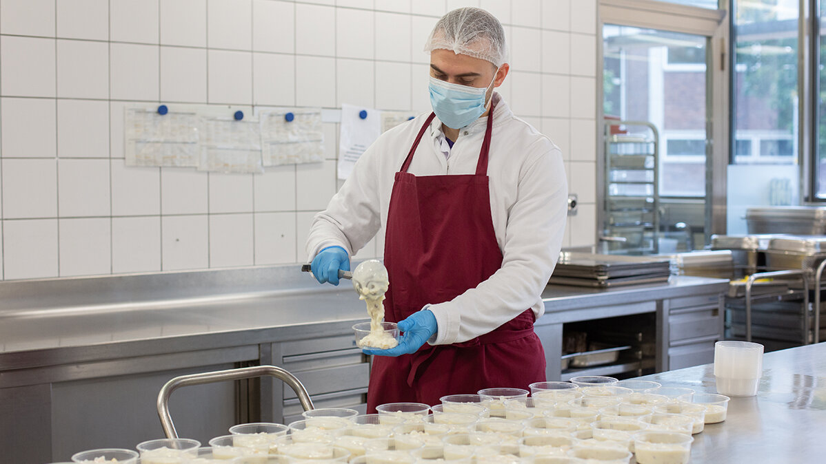 Ruben trägt Haarnetz, Mundschutz und Handschuhe und portioniert in der Küche Kompott.