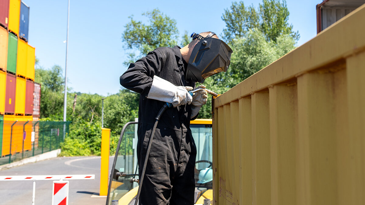 Noah schweißt mit Schutzhelm, Handschuhen und Schutzanzug an einem Container. 