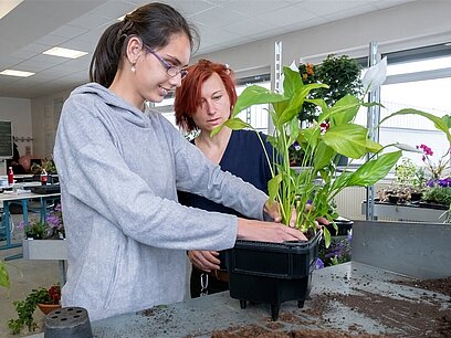 Eine Jugendliche pflanzt in einem Klassenzimmer einer Berufsschule eine Pflanze in einen Blumentopf, während die Ausbilderin daneben steht.