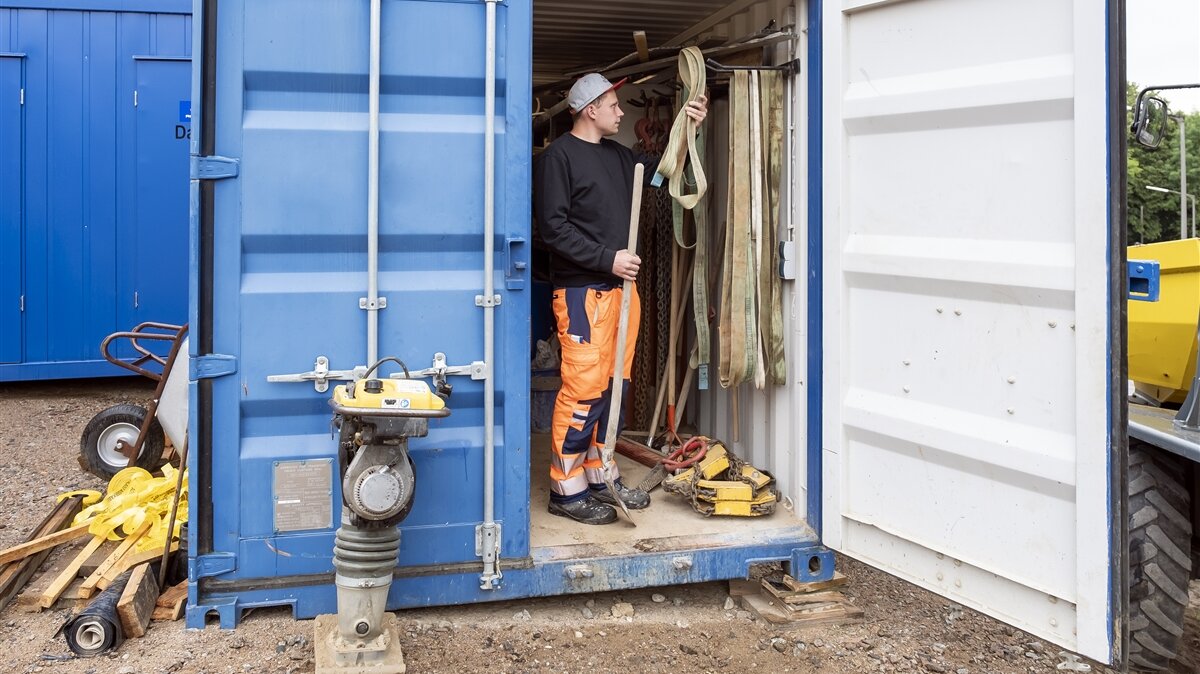 Marius holt auf der Baustelle eine Schaufel und Sicherungsgurte aus dem Werkzeugcontainer.