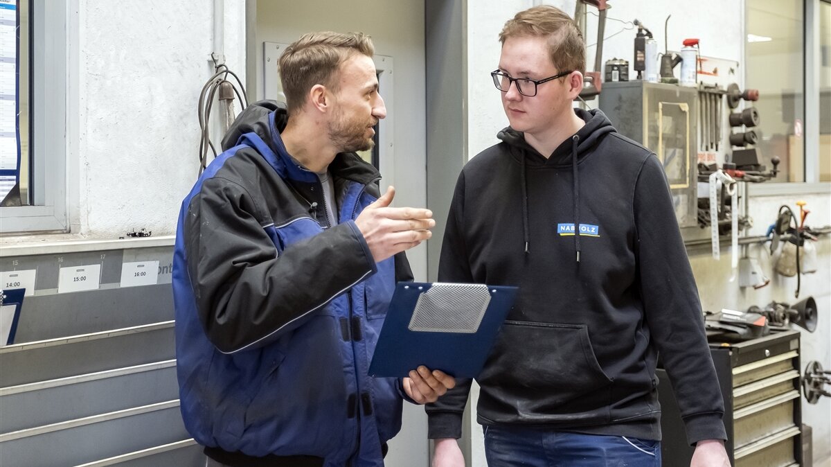Der Ausbilder hält ein Klemmbrett in der Hand und erklärt Tobias seine Aufgabe.