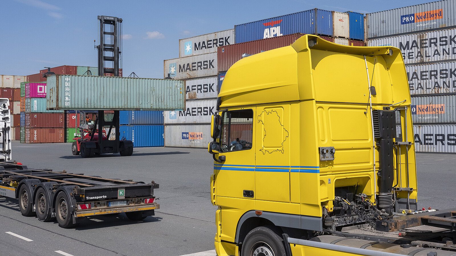 Zwei leere LKWs befinden sich vor dem Verladen von Containern durch einen Gabelstabler.