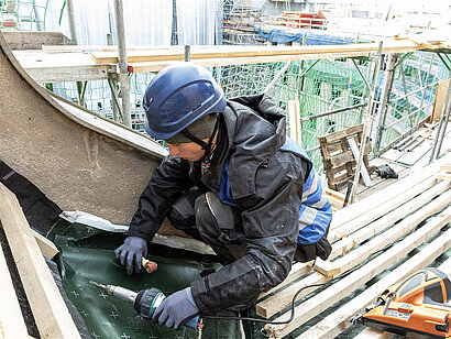 Ein junger Mann mit Helm und Handschuhen schraubt Dämmmaterial an einem Dachstuhl fest.