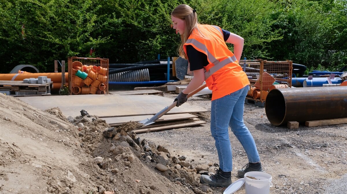 Anna schaufelt mit einem Spaten eine Bodenprobe in einen Eimer.