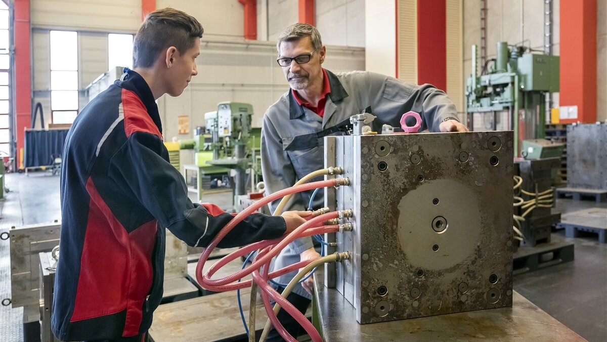 Christian und sein Ausbilder spritzen mit Schläuchen Wasser in die Spritzgussform und prüfen so ob sie dicht ist.