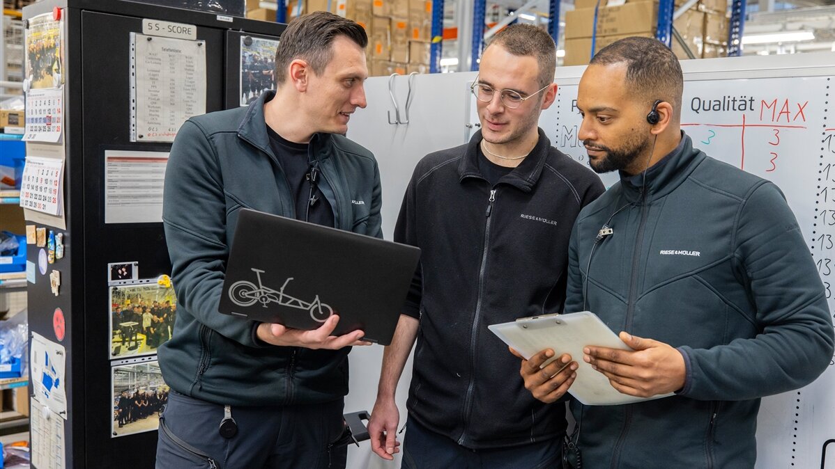 Linus und zwei Kollegen schauen in einer Werkhalle auf einen Laptop.