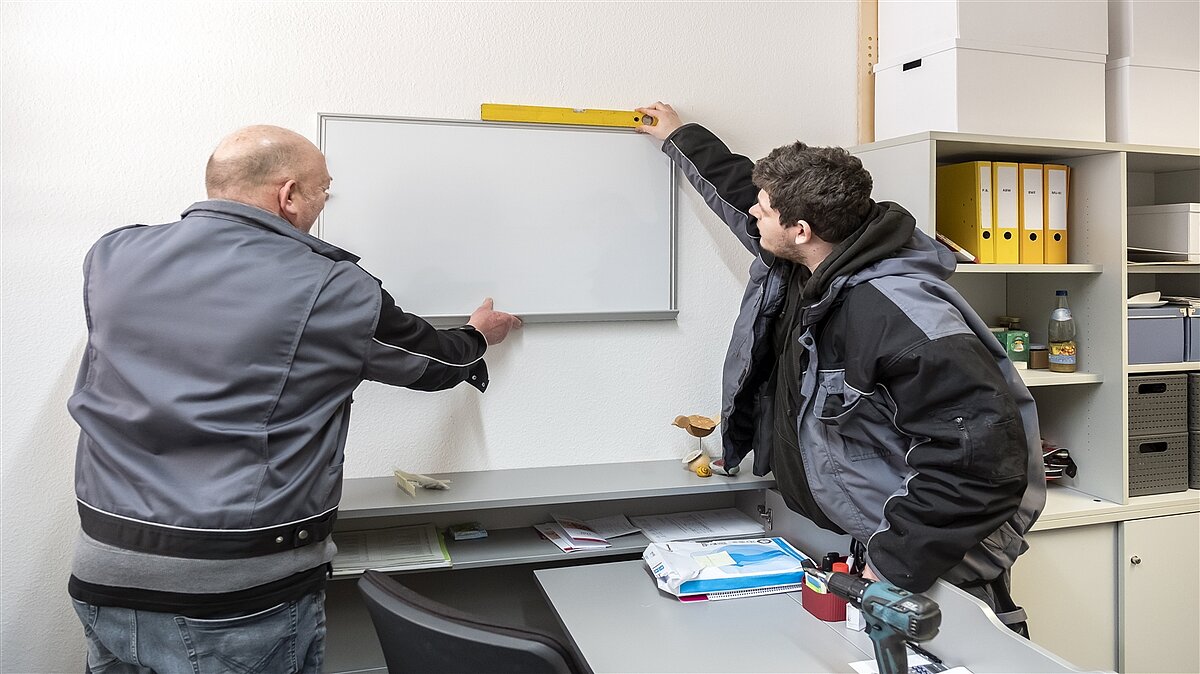 Frank und sein Ausbilder richten das Whiteboard an der Wand aus.