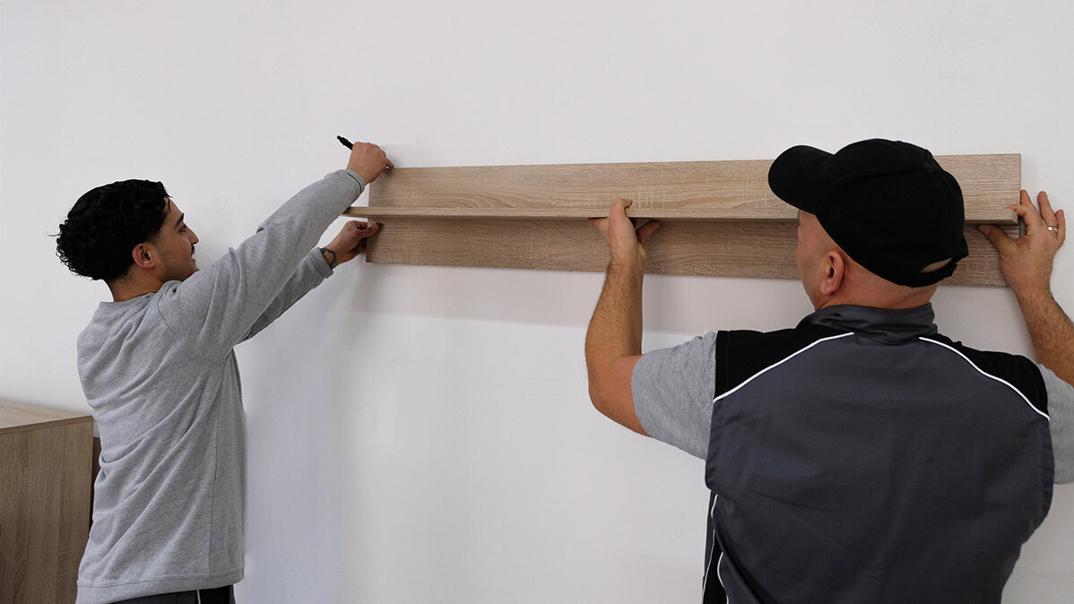 Ein Kollege hilft Zainal das Regalbrett zu halten, damit Zainal die Hilfslinien an der Wand anzeichnen kann.