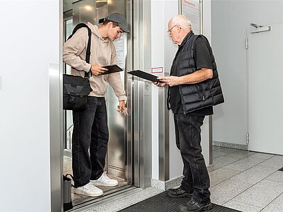 Zwei Männer schauen sich einen Aufzug an und dokumentieren auf einem Klemmbrett und Tablet.